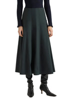 A person stands against a white background wearing the Proenza Schouler White Label Heidi Stretch Cotton Skirt in dark green, styled with a navy textured sweater and black heeled boots.