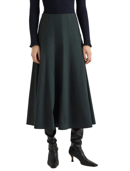 A person stands against a white background wearing the Proenza Schouler White Label Heidi Stretch Cotton Skirt in dark green, styled with a navy textured sweater and black heeled boots.