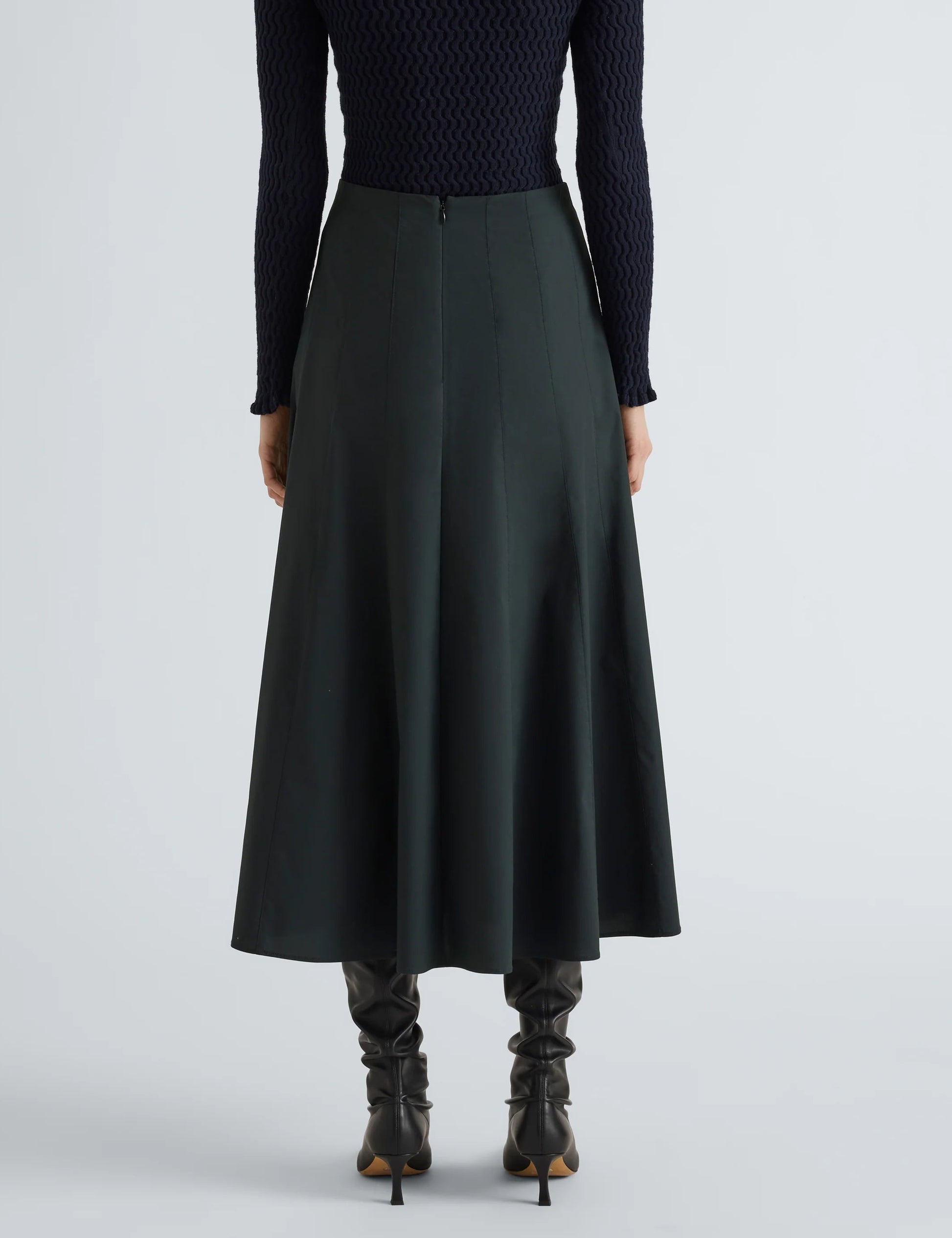 A person is shown from the back against a plain background, wearing the Proenza Schouler White Label Heidi Stretch Cotton Skirt in dark green, styled with a black textured long-sleeve top and black knee-high boots.