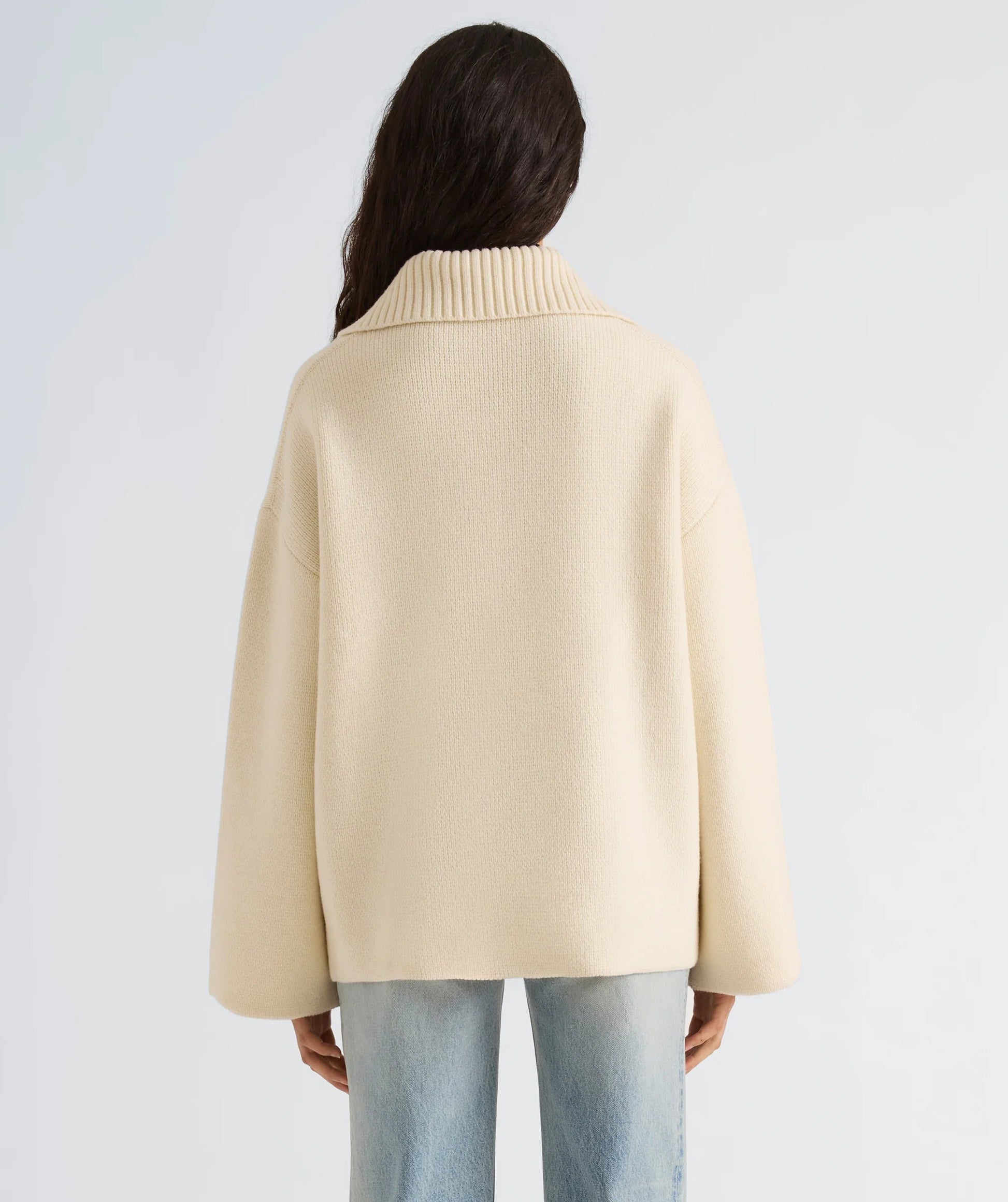 Facing away, the person is wearing the cream-colored, oversized Nelly Melange Merino Jacket by Proenza Schouler White Label, featuring a ribbed collar and paired with light blue jeans against a plain background.