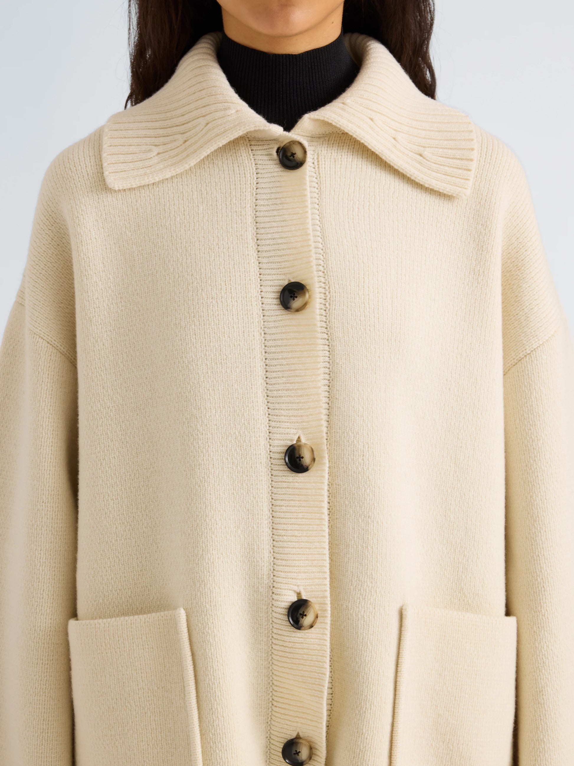 A person wearing the Nelly Melange Merino Jacket by Proenza Schouler White Label, a cream-colored button-up knit cardigan featuring large front pockets and a wide collar, styled over a black turtleneck.