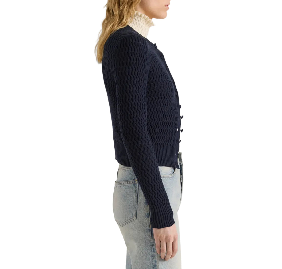 Side view of a person wearing a navy blue Proenza Schouler White Label Marci Cardigan in Ric Rac Cotton over a cream-colored turtleneck and light-wash jeans.