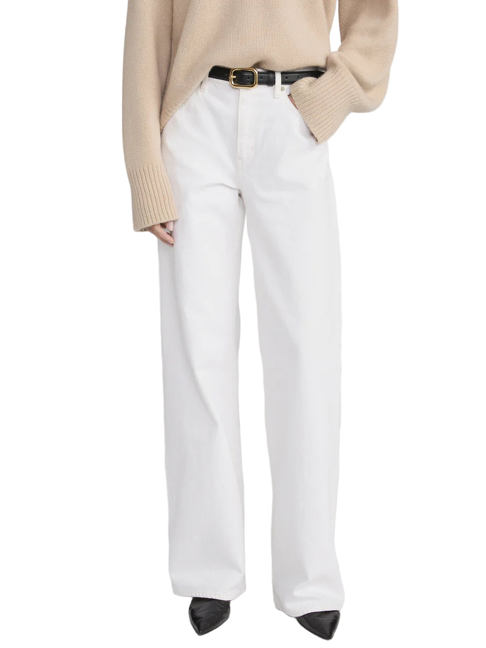 A person wearing The Stroll wide-leg pants by Frame, styled with a beige sweater, black belt, and black pointed shoes, stands with one hand in their pocket.