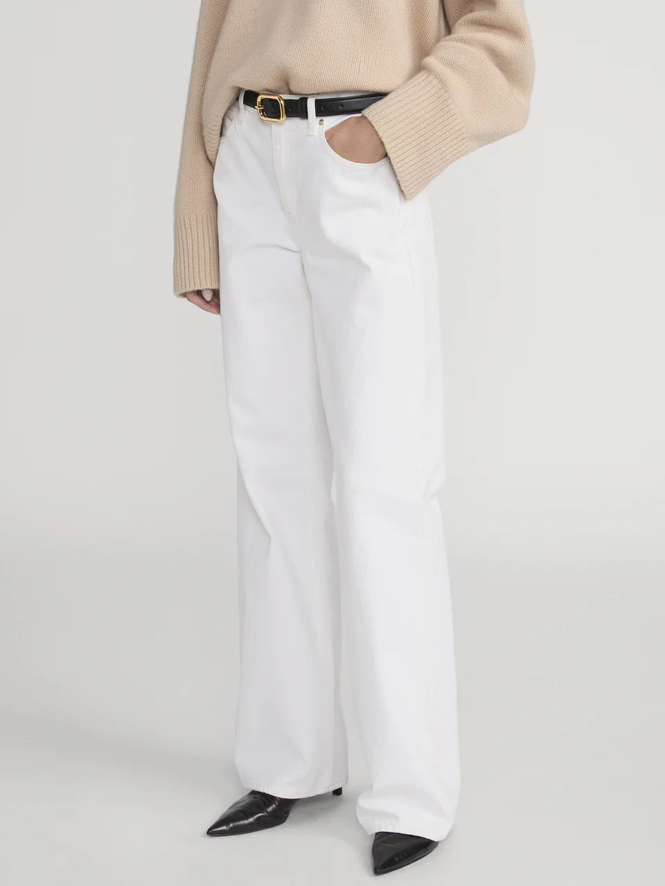 Model wearing The Stroll white mid-rise jeans in premium cotton non-stretch denim by Frame, paired with a beige sweater, black belt, and black pointed shoes, stands with one hand in pocket against a plain background.