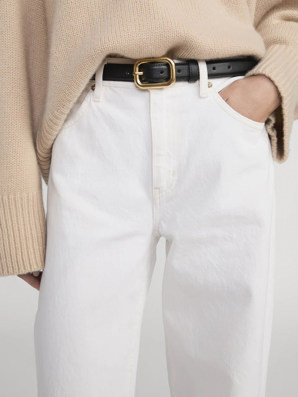 A person wearing Frame's The Stroll beige knit sweater, paired with premium cotton white high-waisted jeans and a black belt with a gold buckle, stands with one hand in their pocket.