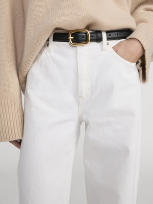 A person wearing Frame's The Stroll beige knit sweater, paired with premium cotton white high-waisted jeans and a black belt with a gold buckle, stands with one hand in their pocket.