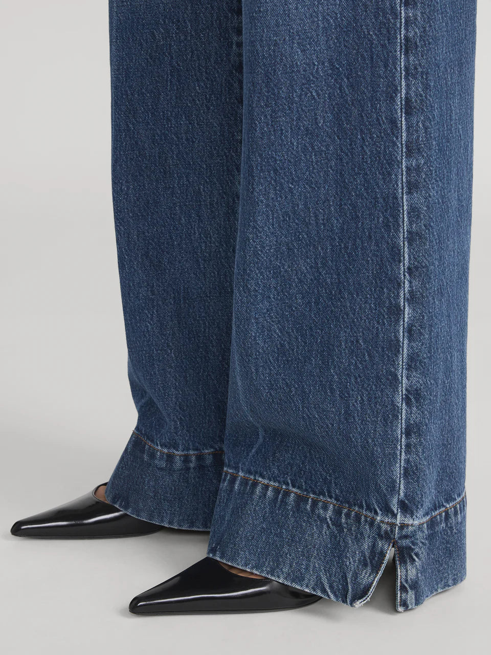 Close-up of a person wearing Frame's The Slice Flare luxury wide-leg blue jeans featuring small hem slits, paired with black pointed-toe shoes.