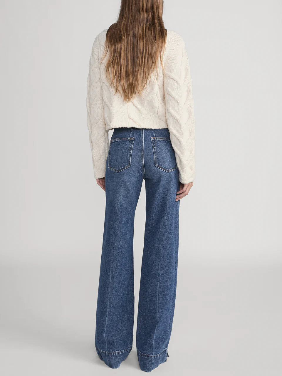 A person stands facing away, wearing Frame’s The Slice Flare ultra-high-rise jeans with frayed hems and a cream cable-knit sweater, set against a plain background.