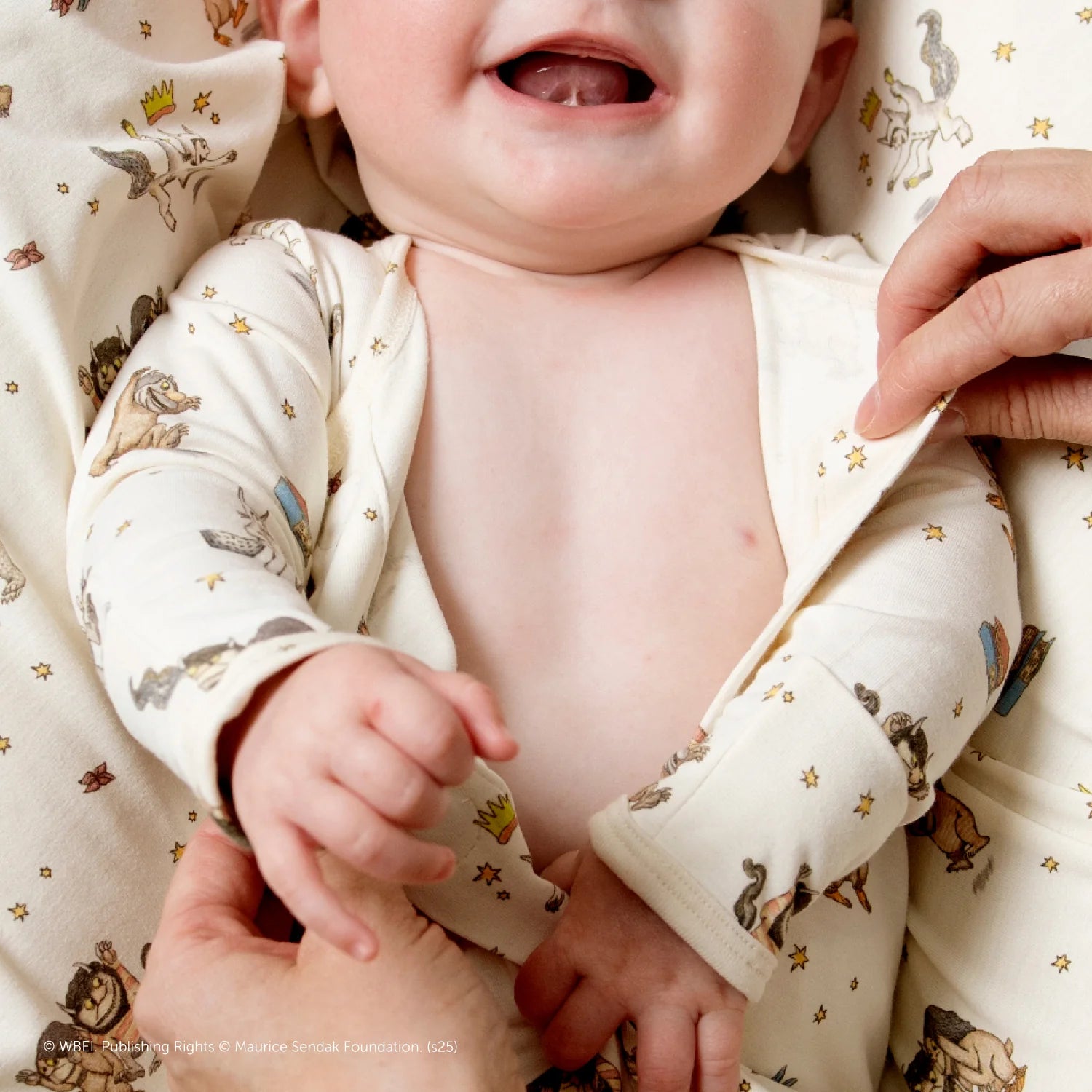 A smiling baby in the Magnetic Me Baby' Where the Wild Things Are Footie, featuring animal and crown designs, has their outfit adjusted by an adult hand. Made from RightFit fabric, these pajamas offer cozy comfort on a light background.