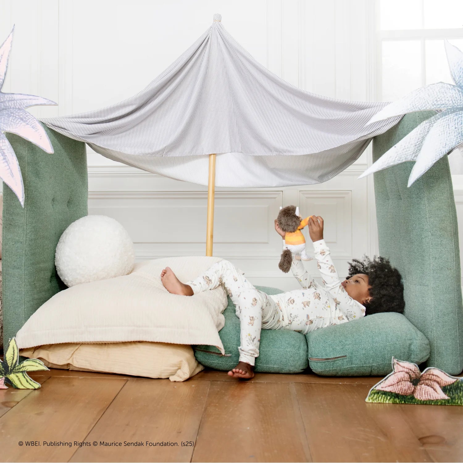 A child in a Magnetic Me Baby and Kids' Where The Wild Things Are PJ Set lies on cushions under a makeshift tent, holding a stuffed animal, surrounded by large illustrated flowers on a wooden floor.