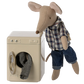 A Maileg mouse in a plaid shirt and jeans stands beside the Maileg Washing Machine, Mouse, with clothes partly inside, part of the adorable mouse family tending to their toy appliances.