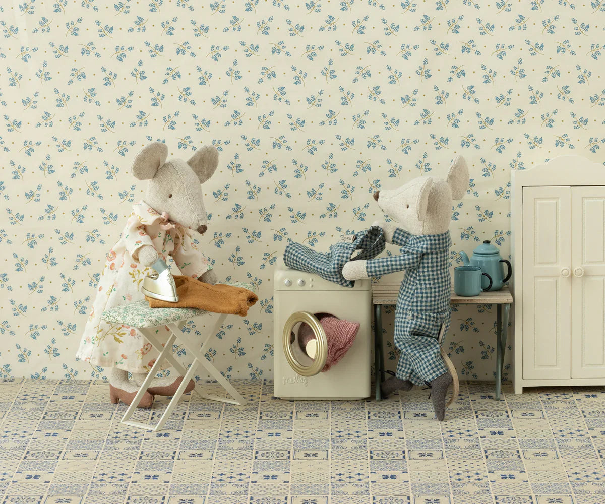 Two fabric mice in a dollhouse setting: one mouse in pajamas stands by a Maileg Washing Machine, Mouse by Maileg, while another mouse in a dress holds an ironing board. The charming scene depicts the mouse family diligently doing laundry.