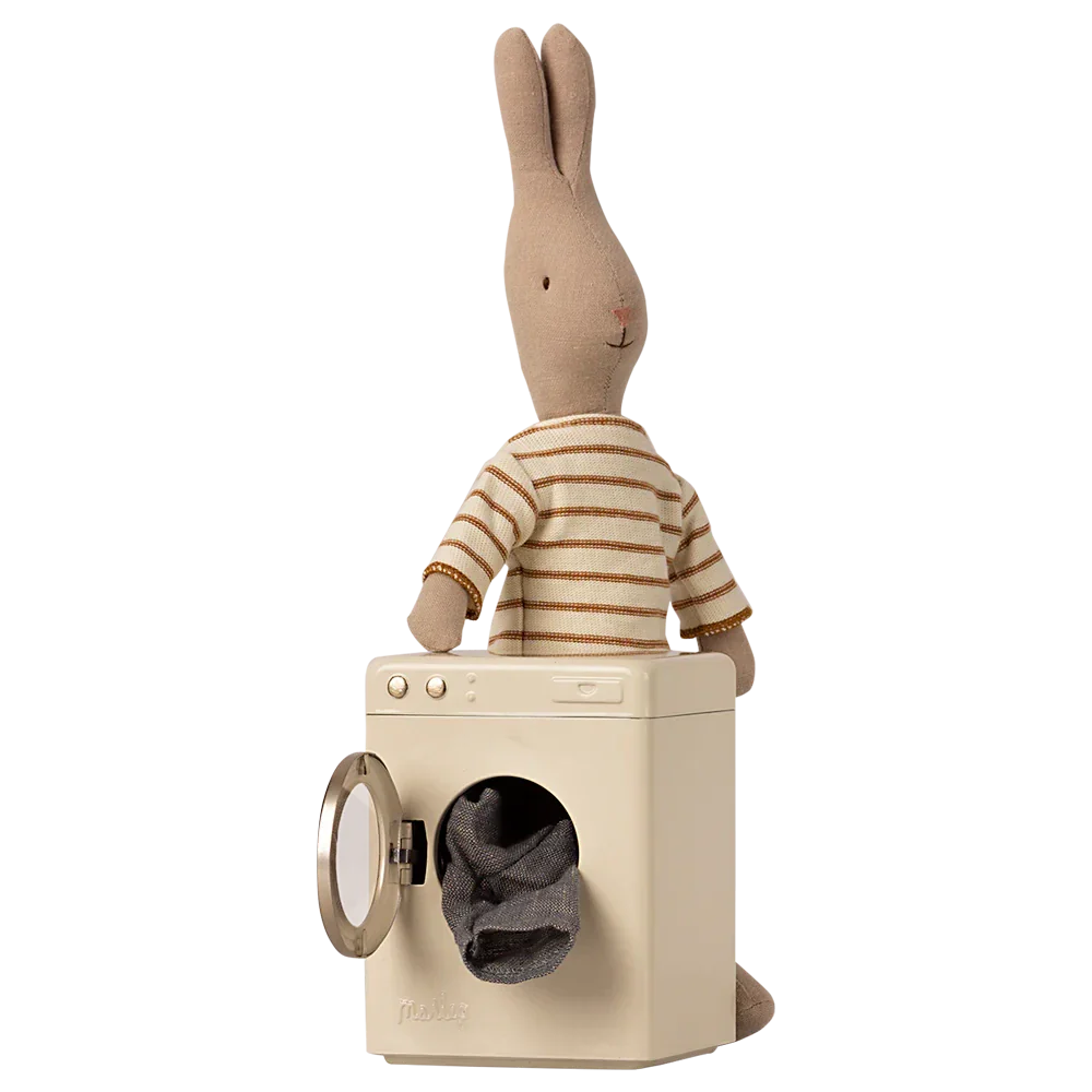 A Maileg Washing Machine, Miniature stands with its door open, showcasing a small laundry load inside, while a stuffed rabbit in a striped shirt sits on top.