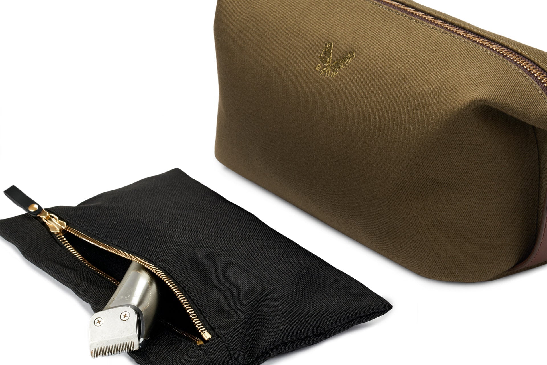 The Bennett Winch Canvas Wash Bag, distinguished by its brown canvas exterior and embroidered logo, is equipped with a durable YKK brass zipper. Next to it, a black zippered pouch holding a silver grooming tool is elegantly displayed on a white surface.