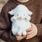 A child in a brown sweater hugs the Wee Kiddo Lamb plush by Bunnies By the Bay, featuring charming small ears and a pink nose, with swirly fur and velour-lined paws for added softness.