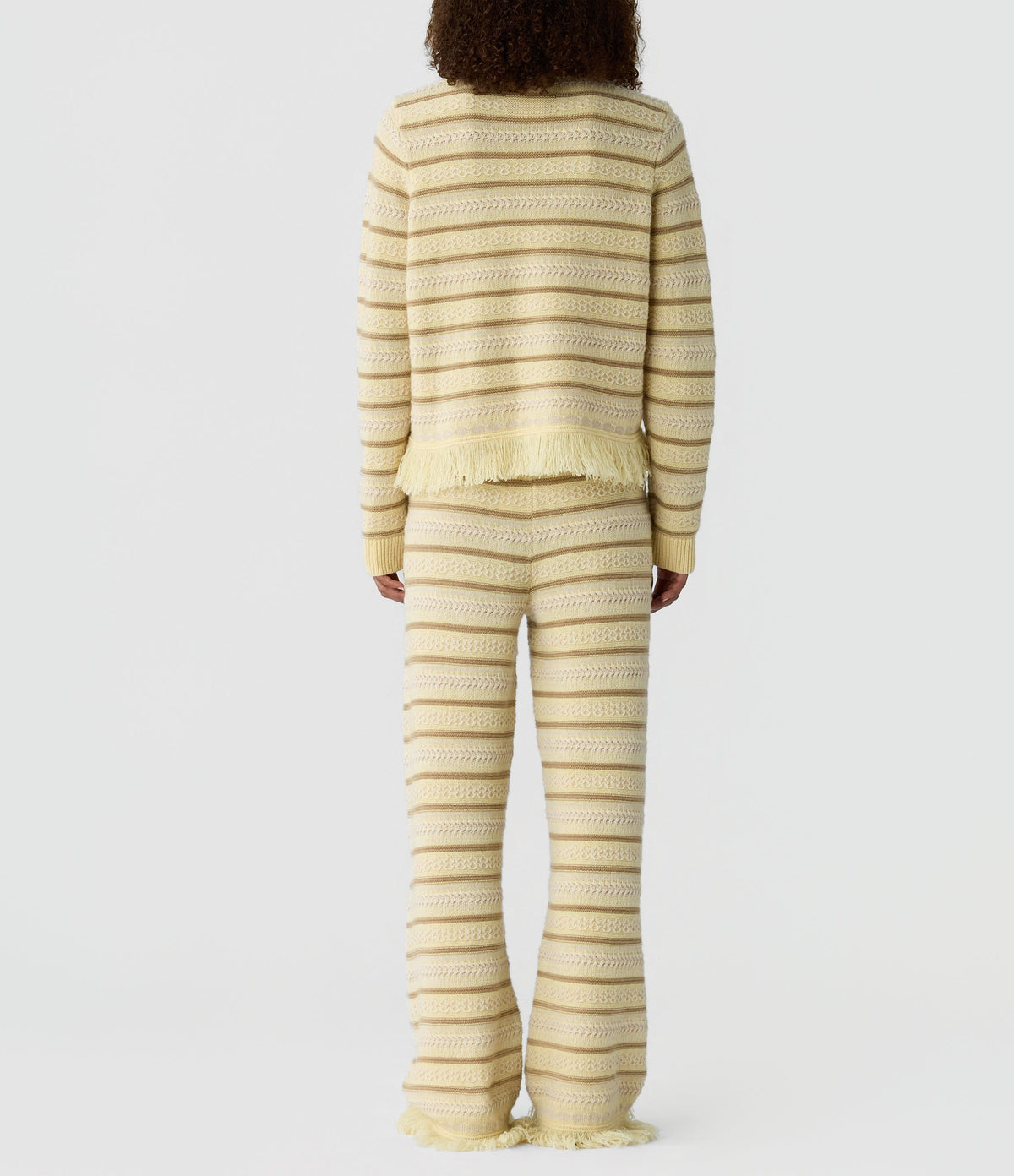 Model wearing the Western Stripe Crew by Guest in Residence, a cream and beige striped knit sweater with a fringed hem, viewed from the back against a white background.