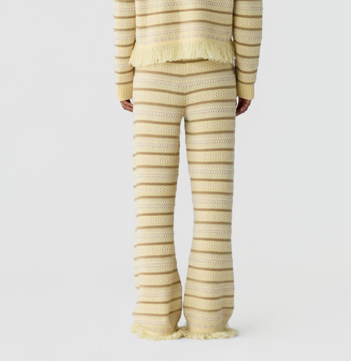 A person stands facing away in a cream and beige knit sweater and the Western Stripe Pant by Guest in Residence, featuring fringe details, against a white background.