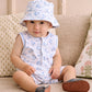 A baby wearing the Kissy Kissy Baby Boys’ Whale Pods Sleeveless Playsuit and a matching bucket hat sits on a beige couch, looking at the camera.
