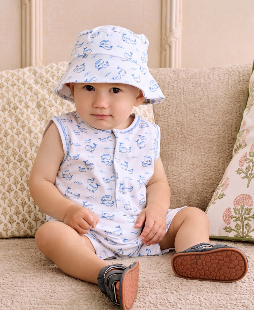 A baby wearing the Kissy Kissy Baby Boys’ Whale Pods Sleeveless Playsuit and a matching bucket hat sits on a beige couch, looking at the camera.