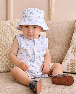 A baby wearing the Kissy Kissy Baby Boys’ Whale Pods Sleeveless Playsuit and a matching bucket hat sits on a beige couch, looking at the camera.