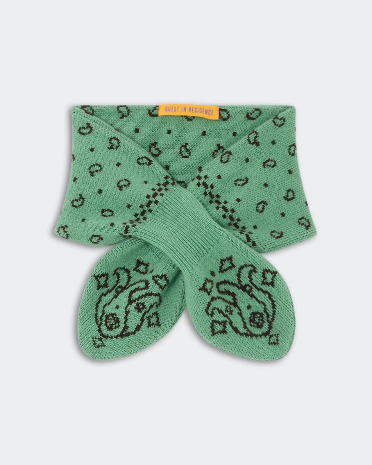 A pair of green socks featuring a black paisley pattern is folded neatly with a yellow “Guest in Residence” tag, paired with the Bandana Bowtie Scarf by Guest in Residence for a coordinated look.