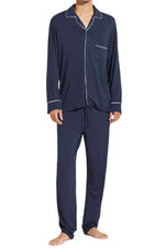 A person wears Eberjey's Willam Long PJ Set, men's dark blue with white piping in sustainable TENCEL™ Modal. Featuring a long-sleeve button-up top and matching pants, these PJs regulate temperature. The barefoot individual stands against a white background.