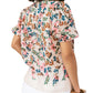 A woman is seen from the back wearing the Figue Wilma Top, a light short-sleeve blouse in airy cotton voile with a pink, blue, and brown floral pattern, pintucked front yoke, split neckline, and paired with white pants.