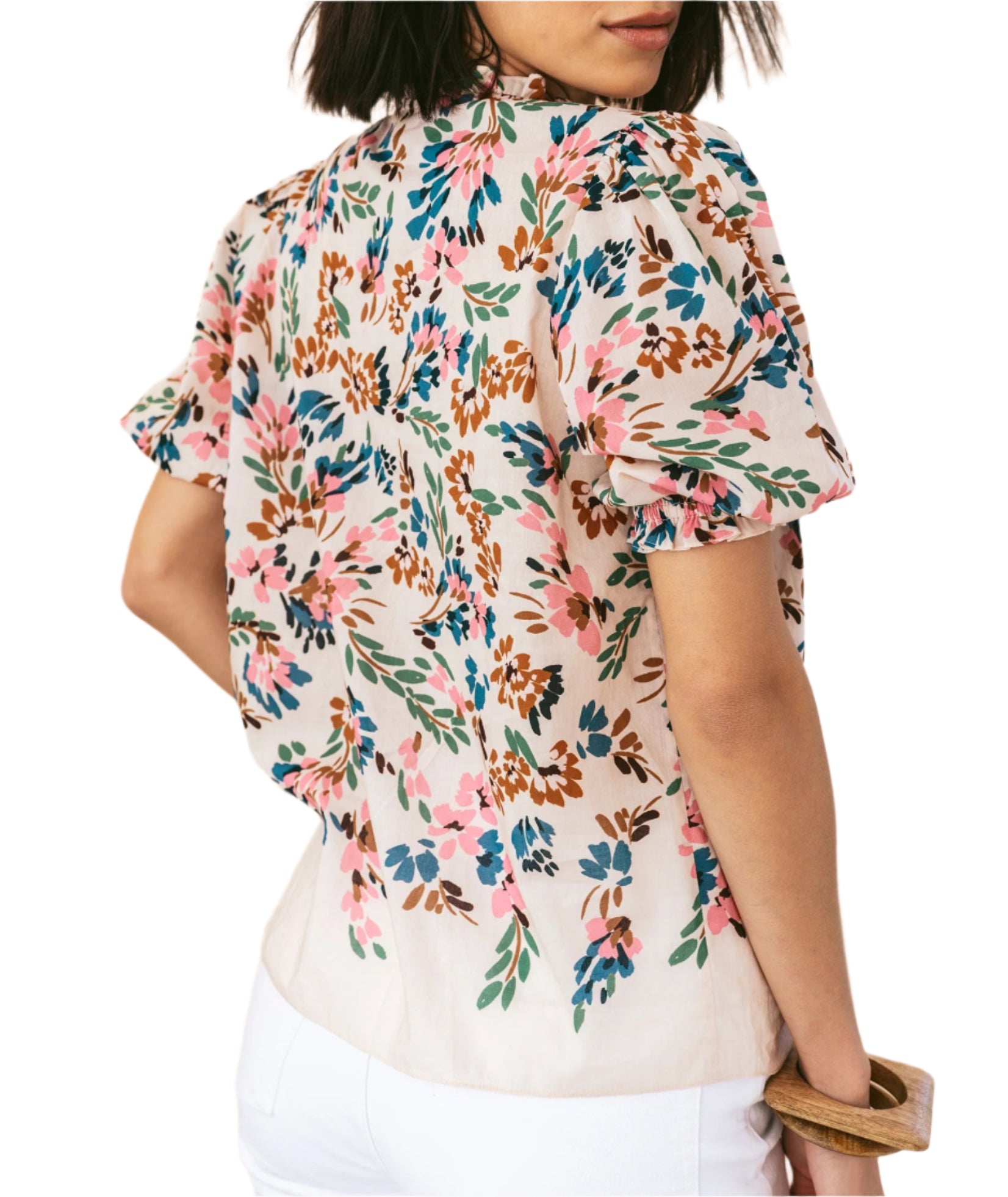 A woman is seen from the back wearing the Figue Wilma Top, a light short-sleeve blouse in airy cotton voile with a pink, blue, and brown floral pattern, pintucked front yoke, split neckline, and paired with white pants.