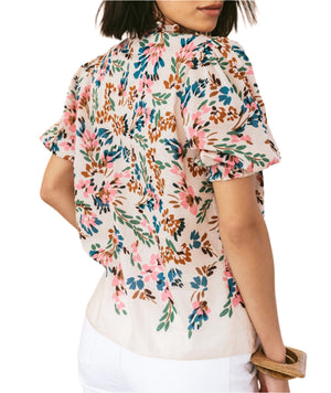 A woman is seen from the back wearing the Figue Wilma Top, a light short-sleeve blouse in airy cotton voile with a pink, blue, and brown floral pattern, pintucked front yoke, split neckline, and paired with white pants.