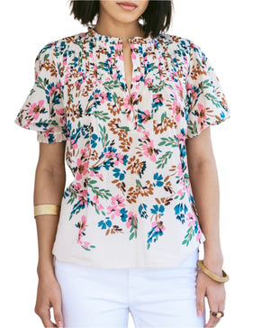 A woman models the Figue Wilma Top, a short-sleeved floral blouse with a split neckline and pintucked yoke in lightweight cotton voile, styled with white pants and gold bracelets.