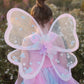 A young child with a bun hairstyle wears a pink and blue kids' costume, complete with the Great Pretenders Twinkling Star Confetti Wings, standing outdoors in a field of flowers.