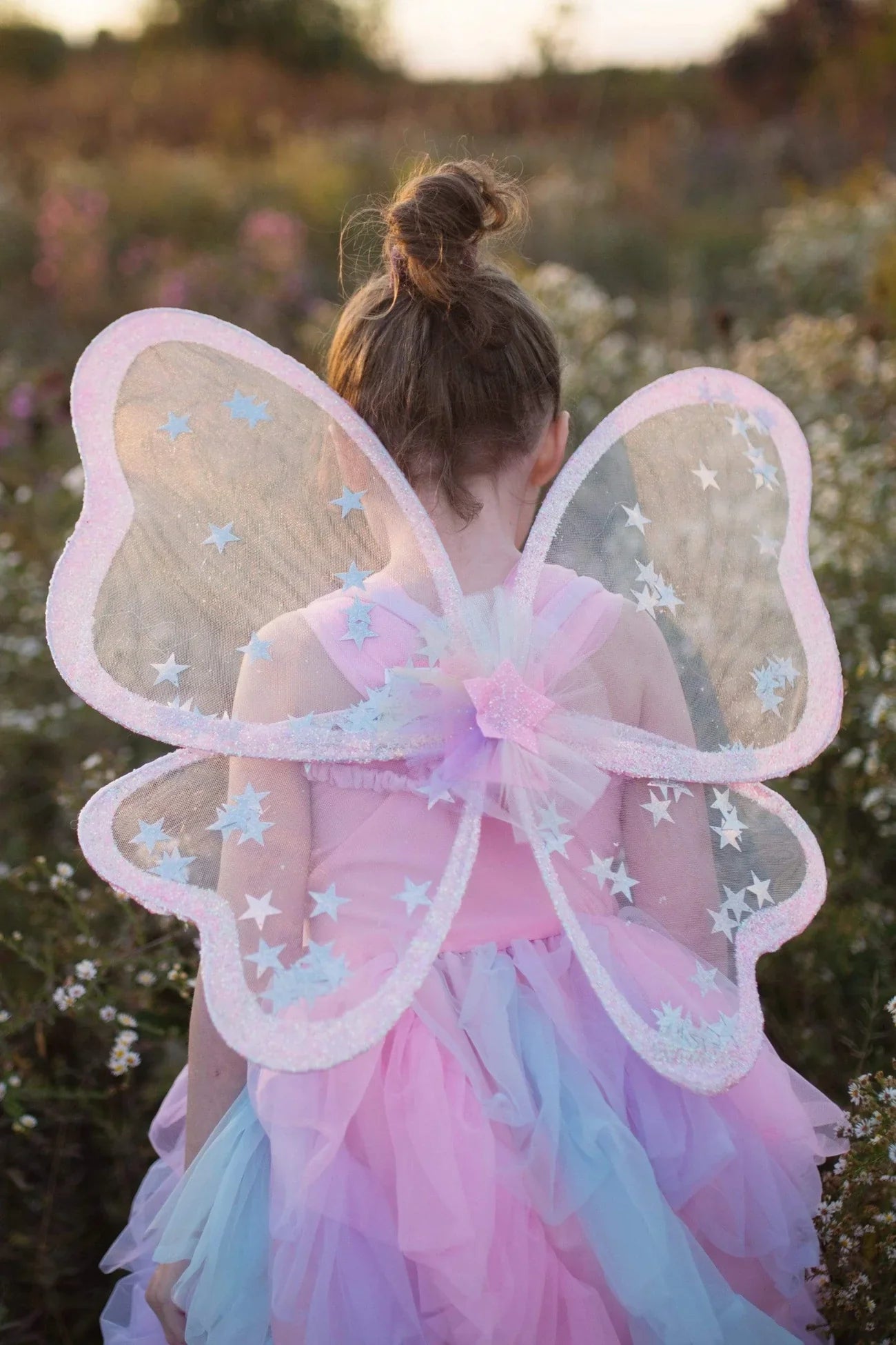 A young child with a bun hairstyle wears a pink and blue kids' costume, complete with the Great Pretenders Twinkling Star Confetti Wings, standing outdoors in a field of flowers.