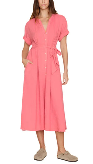 A person wearing the Xirena Cate Dress, a knee-length, short-sleeved pink washed cotton gauze dress with buttons down the front and an adjustable tie-belt at the waist, paired with beige slip-on shoes.