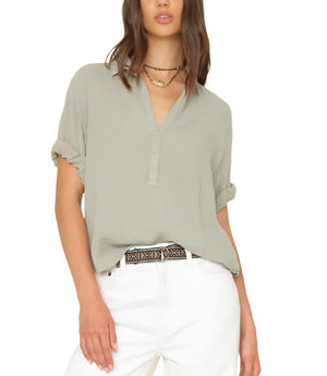 A woman wearing a light green Xirena Cruz Top with rolled-up sleeves, a layered necklace, a patterned belt, and white pants.