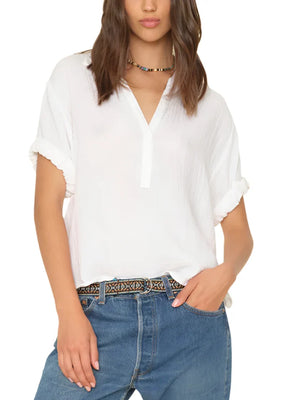 A person wearing the Xirena Cruz Top, a stylish white V-neck henley blouse by Xirena, and blue jeans with a decorative belt stands with one hand in their pocket.