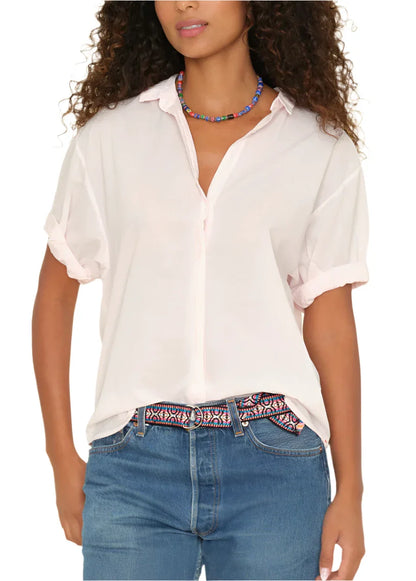 A person with curly hair wears a white Xirena Channing Shirt with rolled sleeves, blue jeans, a colorful beaded necklace, and a patterned belt, exuding a boy meets girl attitude.