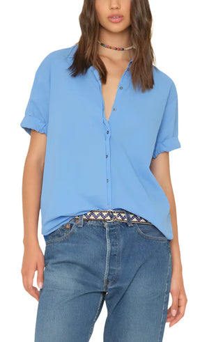 A person wearing a light blue Xirena Channing Shirt made of lightweight cotton poplin, paired with jeans and a beaded necklace, stands against a plain background.