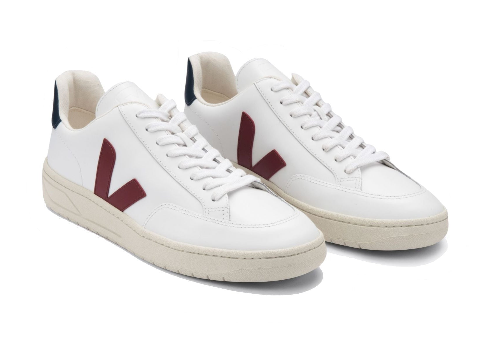 Veja Men's V-12 Leather Sneaker by Veja: Pair of white low-top sneakers with beige soles and maroon "V" logos on the sides. Crafted using recycled materials, the shoes feature white laces, dark blue accents on the heel tabs, and are partly made from organic cotton for added sustainability.