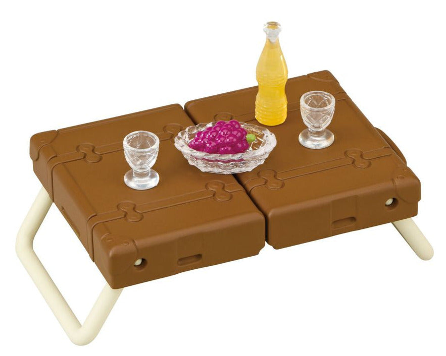 A small brown folding table displays two glasses, a bottle of yellow liquid, a dish of grapes, and Calico Critters Classic Family Car toy accessories arranged neatly on top.