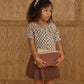 A young girl with curly hair and headband stands holding two books, wearing the Girls' Live Blouse by Rylee & Cru with a scalloped collar and a brown skirt, in front of a wooden paneled wall.