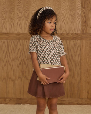 A young girl with curly hair and headband stands holding two books, wearing the Girls' Live Blouse by Rylee & Cru with a scalloped collar and a brown skirt, in front of a wooden paneled wall.
