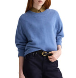 A person wears the TWP Boy Cashmere Crew in blue over an olive green collared shirt, paired with dark pants and a black belt, posing against a white background.