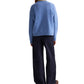 A person stands with their back to the camera, wearing the TWP Boy Cashmere Crew sweater in blue, paired with dark wide-leg pants and brown shoes.