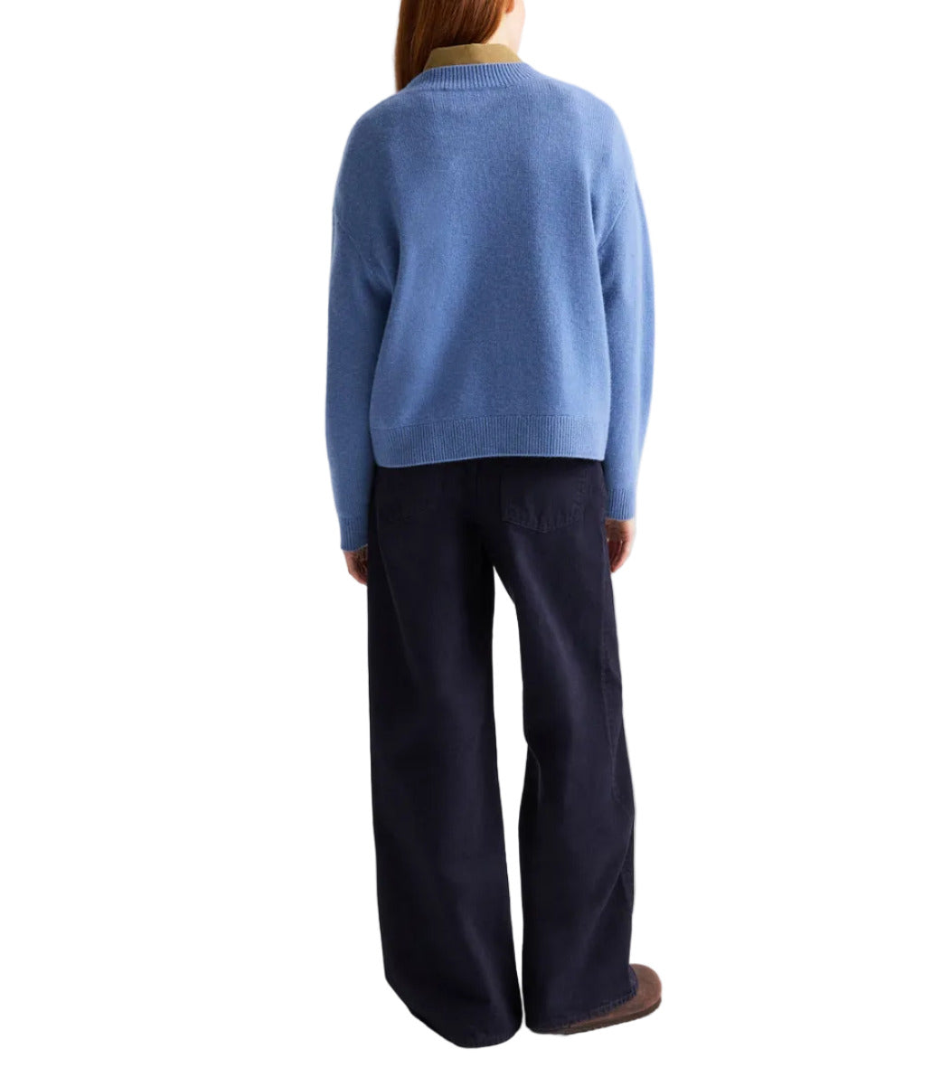 A person stands with their back to the camera, wearing the TWP Boy Cashmere Crew sweater in blue, paired with dark wide-leg pants and brown shoes.