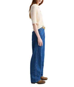 A person stands sideways against a white background, wearing TWP's The Polo knit top in cream with short sleeves and a relaxed fit, paired with wide-leg blue jeans and brown shoes.