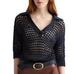 A woman wears the TWP Unspoken Truth Polo, a navy open-knit sweater with a collar and relaxed fit, styled with a black belt featuring a gold buckle and brown pants.