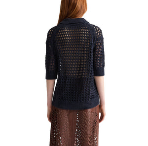 A person with long red hair is shown from the back, wearing TWP’s Unspoken Truth Polo—a dark blue, open-knit sweater with a relaxed silhouette—and a brown, semi-transparent skirt.