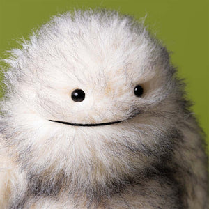 The Jellycat Bretti Yeti, a fluffy white and grey plush toy with black button eyes and a stitched smile, stands as a guardian against a plain green background.