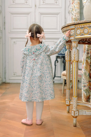 A young girl with two pink bows in her hair stands facing away, wearing the Antoinette Yse Blue Floral Long Sleeve Bishop Dress and white tights, with one hand resting on an ornate table.
