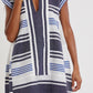 A person wearing the Lemlem Elina Caftan, a short-sleeved, V-neck dress featuring blue, navy, and white stripes with a fringed hem and traditional tibeb weaving inspired by hand-woven Ethiopian designs.